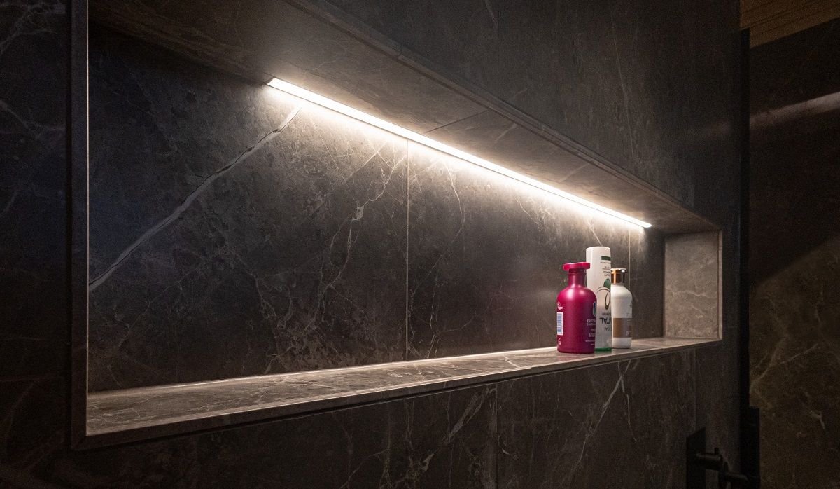 In the dark grey stone wall of a bathroom, the shampoo shelf is illuminated by an LED strip in an aluminium profile with a neutral light colour temperature.